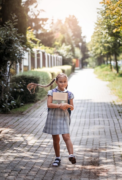 Back to school A cute little schoolgirl in a dress with pigtails and large blue backpackis holds books and walks in the school yard A little girl is going to the first grade