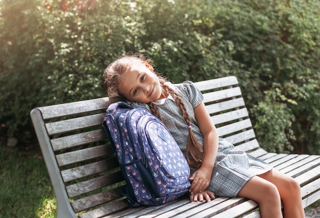 Back to school A cute little schoolgirl in a dress with pigtails and large blue backpackis sitting on a bench in the school yard A little girl is going to the first grade