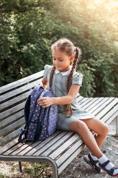 Back to school A cute little schoolgirl in a dress with pigtails and large blue backpackis sitting on a bench in the school yard A little girl is going to the first grade