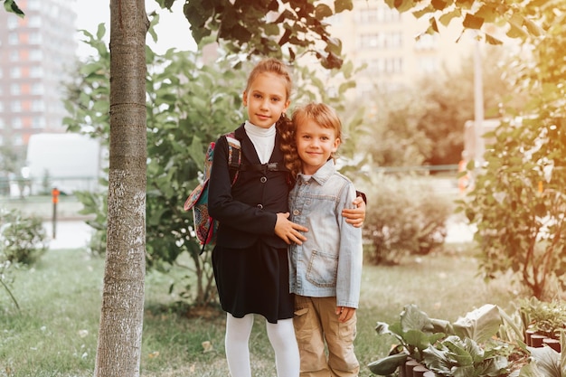 Back to school little happy kid pupil schoolgirl eight years old in fashion uniform with backpack and her preschool brother boy hug together ready going second grade first day primary school flare