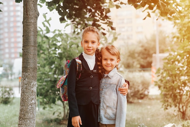 Back to school little happy kid pupil schoolgirl eight years old in fashion uniform with backpack and her preschool brother boy hug together ready going second grade first day primary school flare