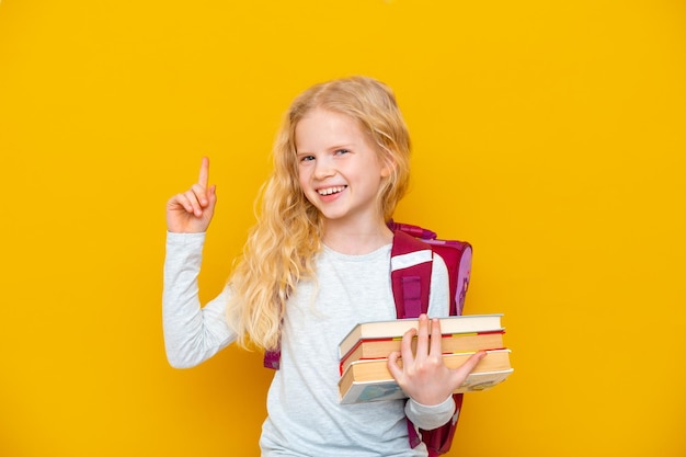 Back to school Portrait of blonde school girl with bag and books Pointing finger up Yellow studio background Education smiling at camera