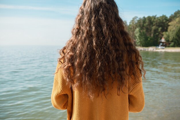 Back shot of female standing on a beach in front of the water in a yellow sweater, selective focus