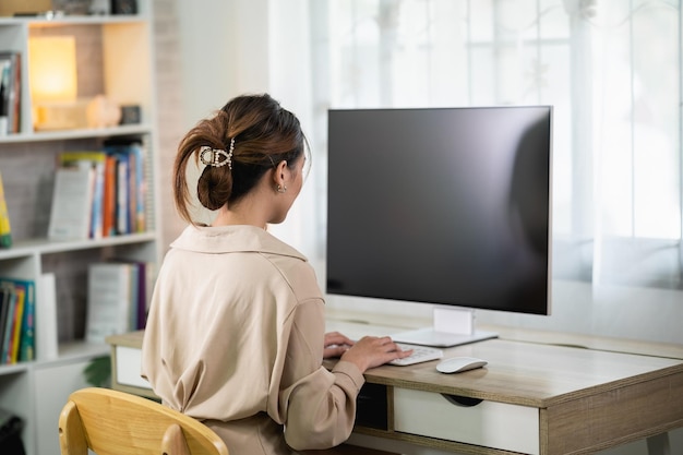 Back side view of Asian freelance woman typing on keyboard and working on laptop on wooden table at home Entrepreneur woman working for her business at living room home Business work home concept