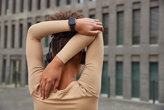 Photo back view of active healthy woman stretches arms warms up before workout wears smartwatch stereo headphones on ears stands against modern city building sports training and wellness concept