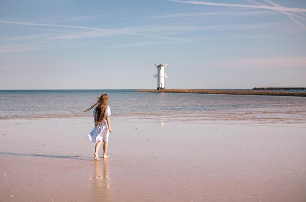 Back view of adorable little girl with long hair in white dress walking on tropical beach vacation