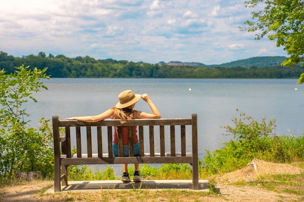 Back view of attractive young woman relaxing on a bench outdoors in front of a lake