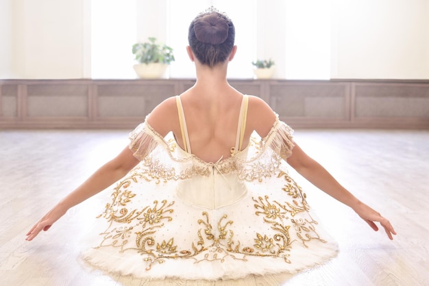 Back view of beautiful ballerina in cream dress sitting and warms up her hands on wooden floor in ballet studio