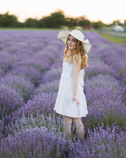 Back view of beautiful girl in a white dress and hat in a lavender field at sunset