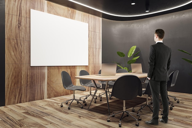 Back view of businessman standing in modern wooden and black meeting room interior with empty white mock up banner equipment furniture and decorative plants Commercial workspace concept