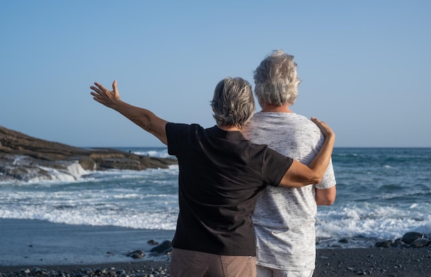 Back view of caucasian senior couple standing on the beach at sunset looking at horizon over water relaxed pensioners enjoying free time sea vacation or retirement