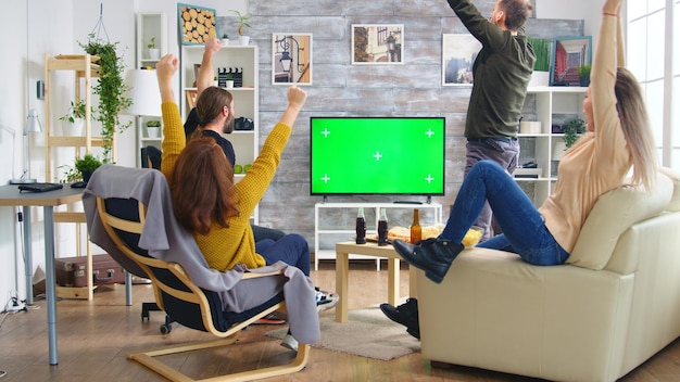 Back view of close friends with hands in the air while watching a football match in living room.