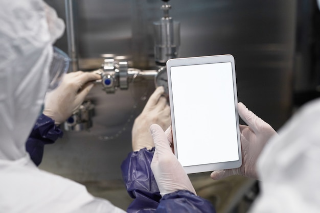 Photo back view close up of two workers holding tablet with blank white screen at modern chemical plant, copy space