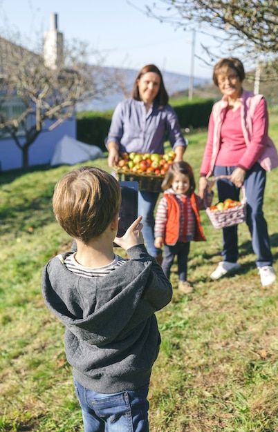 Back view of cute boy taking a photo with electronic tablet to family with fresh organic apples in a wicker basket after harvest. Three different generations concept.