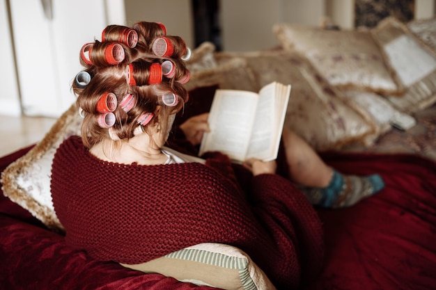 Back view of elderly woman applying colorful haircurlers wearing vinous cardigan sitting on sofa reading book