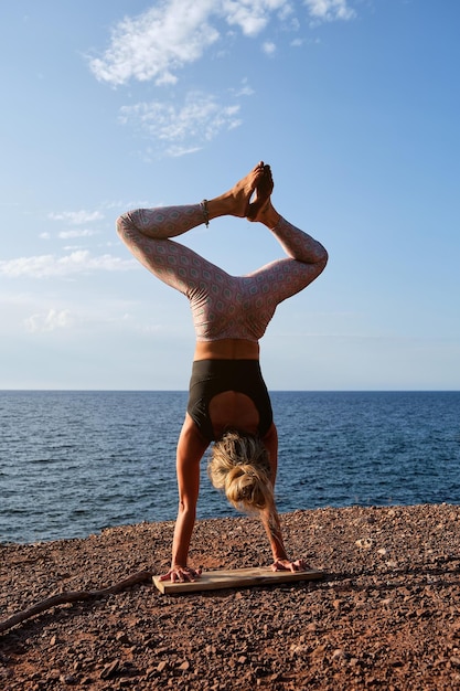 Back view of faceless female athlete in sportswear standing on wooden plank in Adho Mukha Vrikshasana pose with legs in Baddha Konasana while practicing yoga on beach