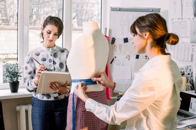 Back view of female dressmaker measuring mannequin with tape in atelier office. Talented fashion shop owner designer working with sketches for new collection of clothes. Dressmaking and sewing concept