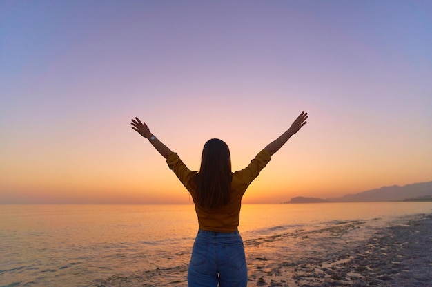 Back view of free calm bliss satisfied woman standing with open arms against of sunset sky by the sea in a happy beautiful inspired moment of her life