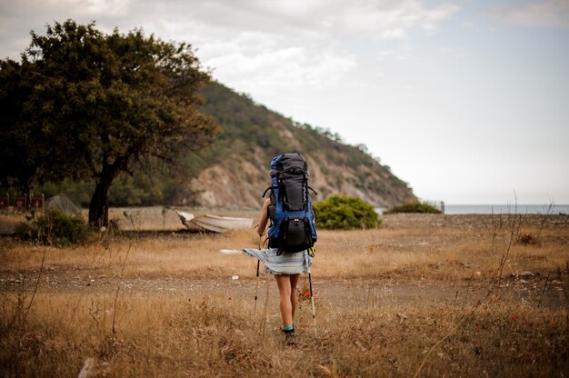 Back view girl with backpack and hiking sticks walking through the field