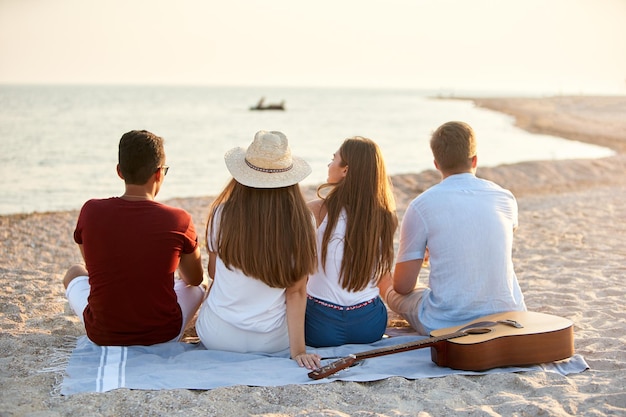 Back view of group of friends sitting together on towel on white sand beach during their vacation and enjoying a sunset above the sea Men and women relax on the shore with guitar Tranquil scene