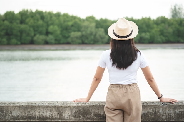 Back view of happy hipster woman in hat standing and looking at river in evening sunset.