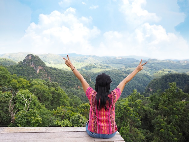 Back view of happy woman tourist raise arms over rainforest mountain and blue sky landscape background.