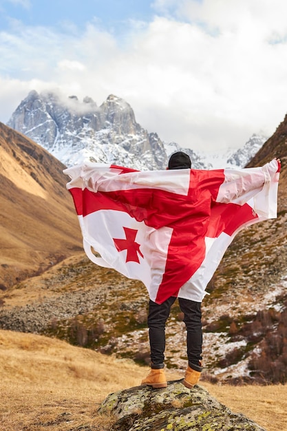 Back view of happy young man posing with georgia national flag standing outdoors looks at snow cappe...