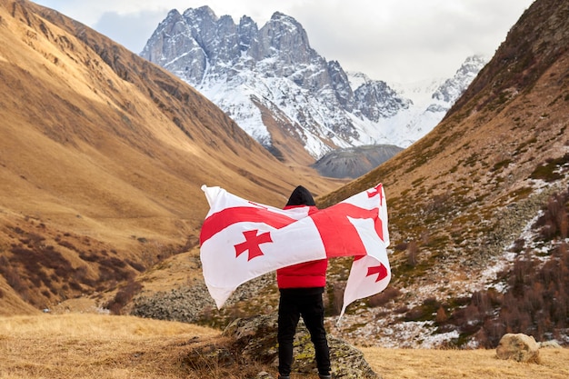 Back view of happy young man posing with georgia national flag standing outdoors looks at snow cappe...