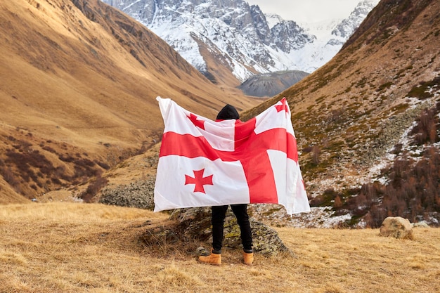 Back view of happy young man posing with georgia national flag standing outdoors looks at snow cappe...