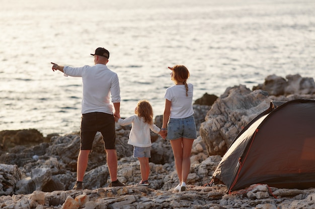 Back view of happy young parents, holding hands daughter, standing on rock beach.