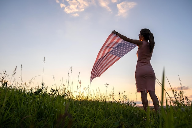 Back view of happy young woman posing with USA national flag standing outdoors at sunset Positive female celebrating United States independence day International day of democracy concept