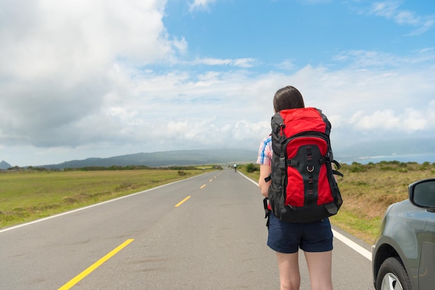 back view of leisurely young female backpacker parking car on asphalt roadside looking at distant mountain scenery ready to hiking in road trip.