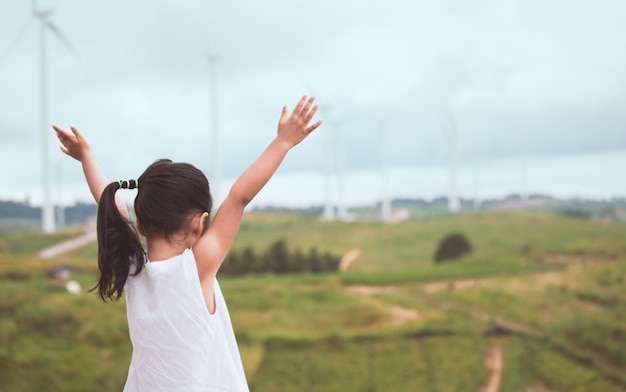 Back view of little asian child girl raise her arms looking at wind turbine field