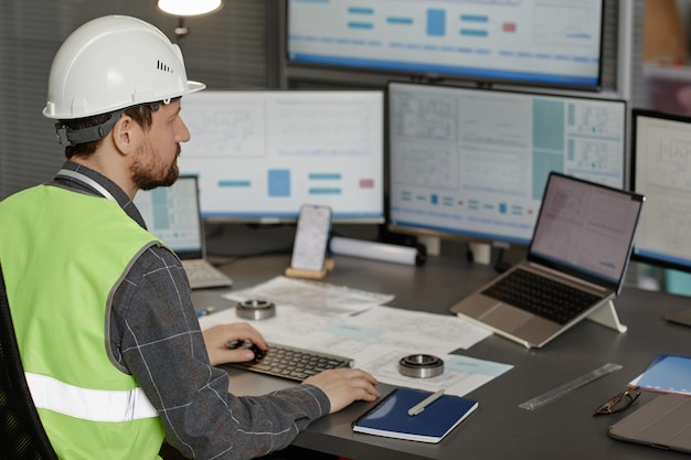 Back view of male construction engineer wearing hardhat at workplace in office and using computer copy space