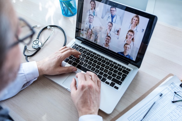 Back view of male doctor having video conference on laptop with his colleagues at home.