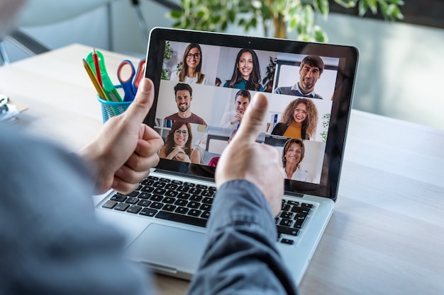 Back view of male employee making a gesturing approval with thumb fingers while speak on video call with diverse colleagues on online briefing with laptop at home.