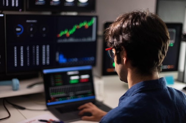 Back view of man in blue shirt working with multiple screens and laptop Stock market and trading