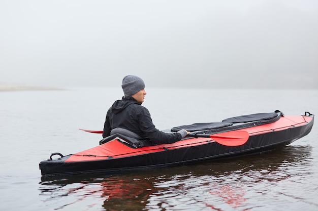 Back view of man paddling kayak on river or lake, looking at nature, wearing black jacket and gray cap, foggy day in open air