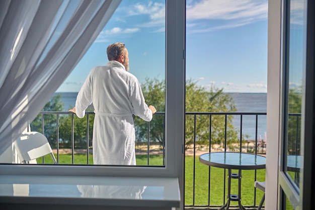 Back view of a man in the white terry bathrobe leaning on the balcony railing