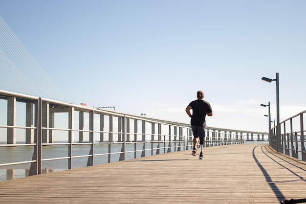 Back view of man with artificial leg jogging on bridge. Sporty man in casual clothes training outdoors in morning running on bridge over sea. Health care, active life of people with disability concept