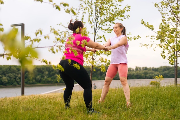 Back view of professional fitness female trainer giving personal training to overweight young woman outdoor in summer day Instructor help fat woman lose weight outside doing squats outside