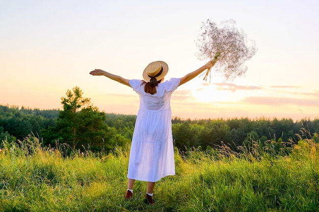 Back view of romantic woman in a white dress with a straw hat enjoying sunset