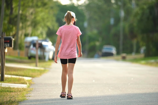 Back view of sad young child girl walking alone along the green street on sunny summer day