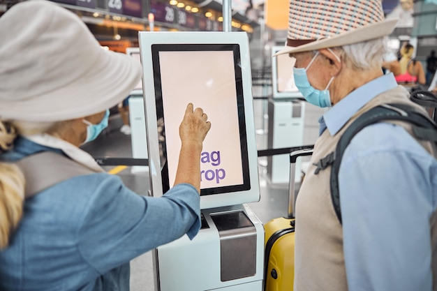 Photo back view of a senior caucasian couple standing before the self-service check-in touch screen kiosk