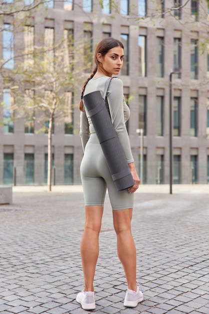 Back view of sporty muscular brunette woman dressed in sportsclothes carries rolled karemat dressed in sportsclothes and sneakers turns behind returs after aerobics or gymnastics stands outdoors