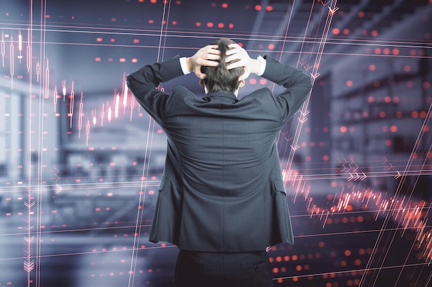 Back view of stressed young european businessman standing in blurry office interior with falling red index candlestick graph Crisis market fall recession and money loss concept Double exposure