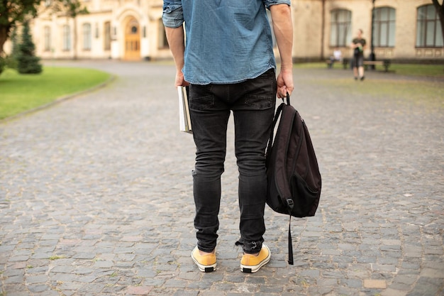 Back view of stylish man in yellow sneakers, denim shirt, black jeans stand with book, waiting.