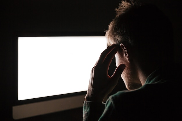 Back view of thoughtful young man thinking and using blank screen computer in the dark room