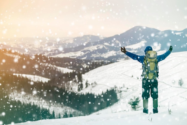 Back view of tourist hiker in warm clothing with backpack standing with raised arms on mountain clearing on copy space background of woody mountain ridge and cloudy sky