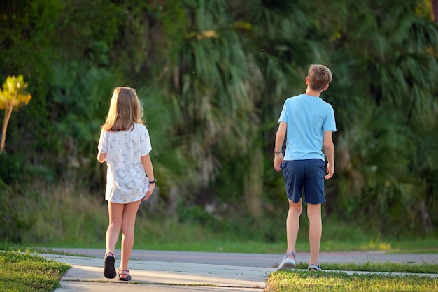 Back view of two young teenage children girl and boy brother and sister walking together on suburban street on bright sunny evening